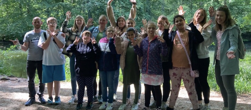 Eine Gruppe von Kindern steht mit ihren Lehrerinnen im Wald - alle winken lachend in die Kamera