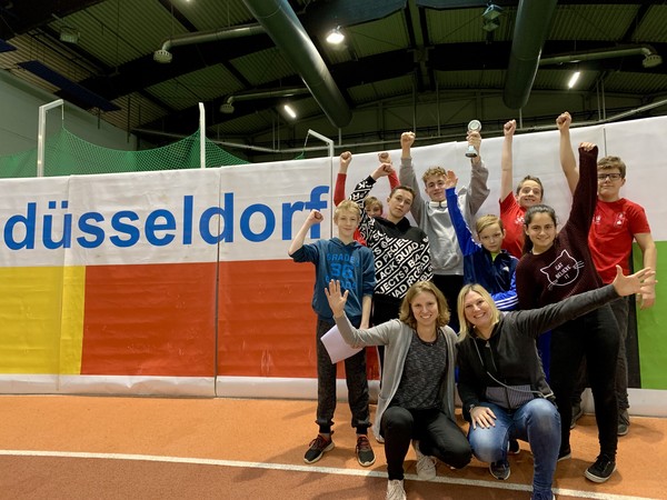 Mehrere Schüler:innen sowie zwei Lehrerinnen stehen für ein Gruppenfoto in einer Sporthalle zusammen und bejubeln ihr Leistung beim Leichtathletik