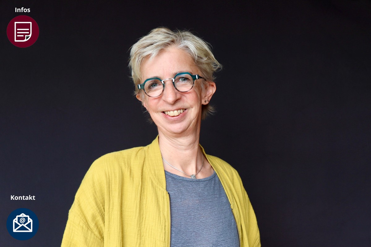 Kurze blonde Haare, eine auffällige türkisfarbene Brille und eine senfgelbe Strickjacke über einem grauen T-Shirt.