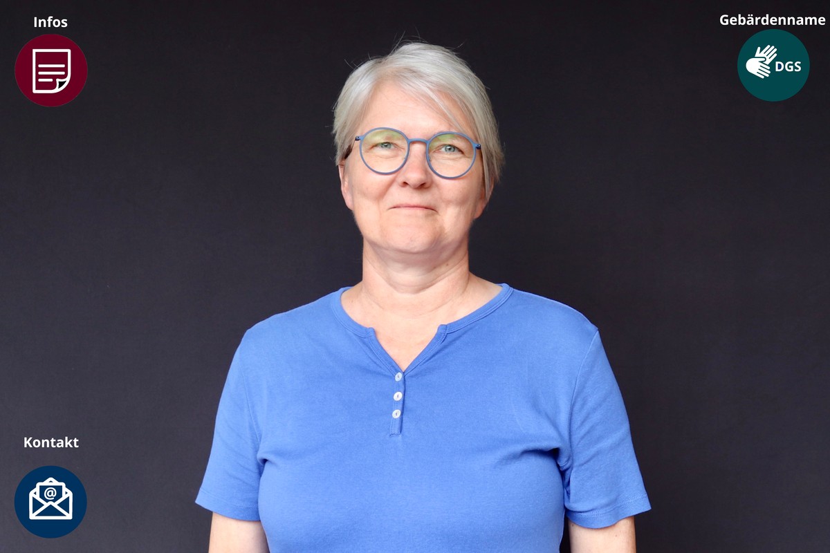 Sehr kurze silberweiße Haare, eine runde türkisblaue Brille und ein leuchtend blaues Shirt mit kleiner Knopfleiste.