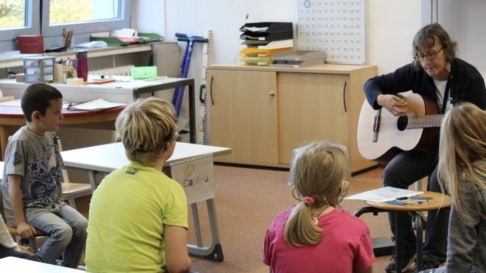Schüler im Alter von 7-8 sitzen an ihren Tischen im Kreis und schauen nach vorne zur mit Gitarre musizierenden Lehrkraft