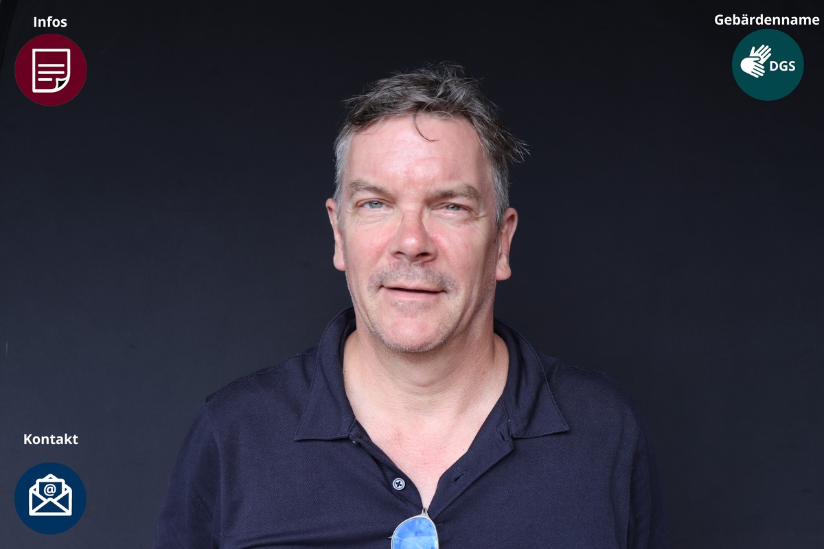 Kurze dunkelbraune Haare, ein Schnurrbart und ein dunkelblaues Poloshirt; eine blaue Brille hängt am Kragen.