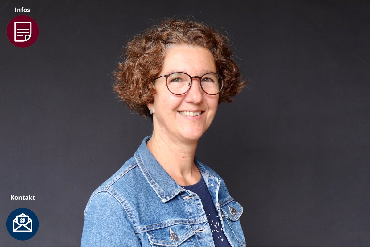 Kurze, voluminöse braune Locken, eine runde dunkle Brille und eine hellblaue Jeansjacke über einem dunkel gemusterten Oberteil.