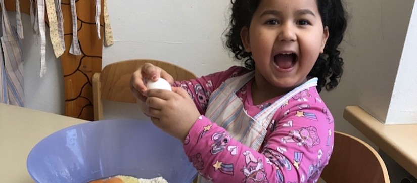 A girl cracks an egg over a bowl and looks at the camera, beaming with joy.