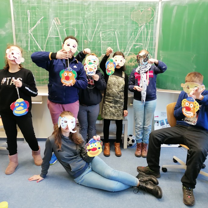 Schüler:innen stehen mit ihren Holz-Clownsköpfen vor einer Tafel. (vergrößerte Bildansicht wird geöffnet)