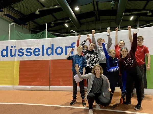 Mehrere Schüler:innen sowie eine Lehrerin stehen für ein Gruppenfoto in einer Sporthalle zusammen und bejubeln ihre Leistung beim Leichtathletik,.